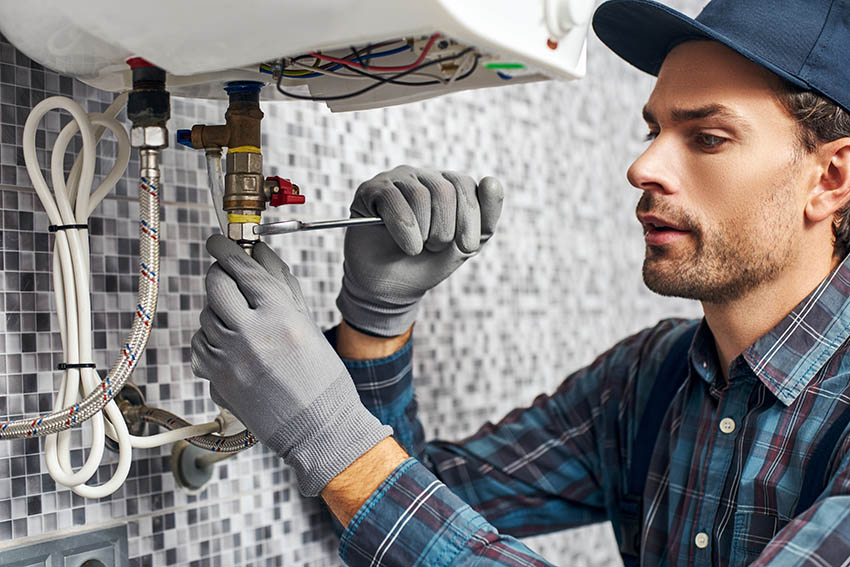 Young plumber look under sink in kitchen. He put new hose. Tools with opened box on floor. Hose and wrench with nippers on desk