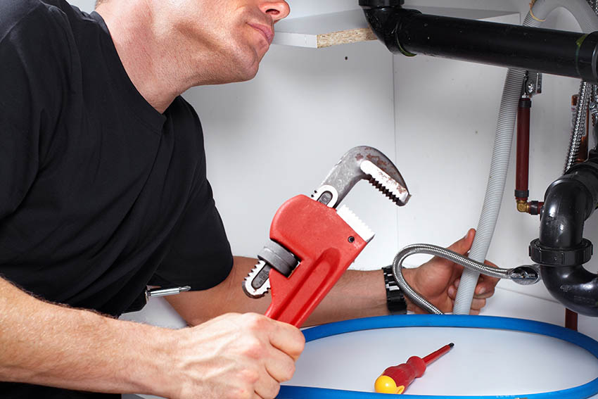 Young plumber look under sink in kitchen. He put new hose. Tools with opened box on floor. Hose and wrench with nippers on desk